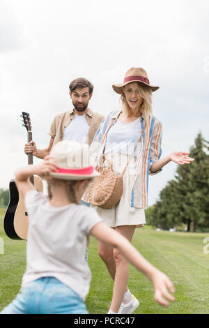 Bella madre e padre felice con la chitarra acustica guardando carino piccola figlia divertirsi nel parco Foto Stock