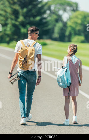 Vista posteriore della ragazza adolescente e ragazzo con zaini, libri e lo skateboard camminare e parlare insieme in posizione di parcheggio Foto Stock