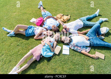 Angolo di alta vista sorridente compagni adolescenti con libri e zaini giacente insieme sul prato verde Foto Stock