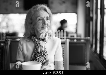 Ritratto in bianco e nero di Senior donna anziana a bere caffè e guardando fuori della finestra Foto Stock
