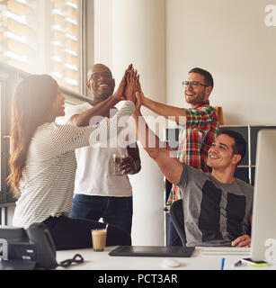 Il gruppo di quattro diversi uomini e donne in abbigliamento casual a mettere i loro palmi insieme in ufficio vicino alla scrivania e grande finestra luminosa Foto Stock
