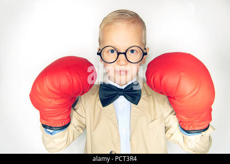 Carino piccolo ragazzo in un vestito e cravatta a farfalla oversize da indossare guanti da boxe e guardando intrepido mentre in piedi contro uno sfondo bianco Foto Stock