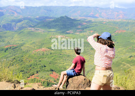 Un giovane maschio si siede su una roccia mentre una giovane donna in piedi dietro di lui le fotografie del Canyon di Waimea in Kauai, Hawaii, Stati Uniti. Foto Stock
