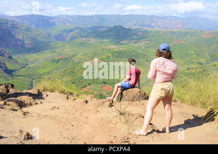 Un giovane maschio si siede su una roccia e una giovane donna sta dietro di lui come essi contemplano il Canyon di Waimea in Kauai, Hawaii, Stati Uniti. Foto Stock
