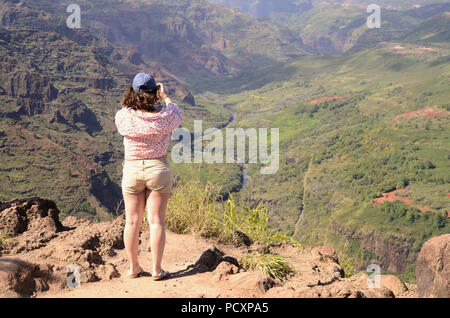 Giovane donna con il suo ritorno verso la fotocamera per scattare una foto sul suo telefono cel del Canyon di Waimea in Kauai, Hawaii, Stati Uniti. Foto Stock