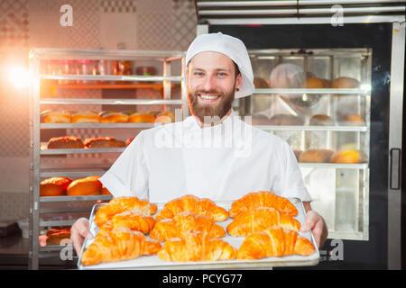 Giovane bello baker holding croissant appena sfornati in mani sullo sfondo del forno. Foto Stock