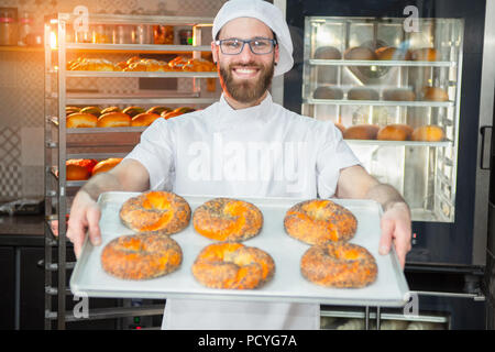 Un giovane bello baker holding bagel freschi con semi di papavero su un vassoio sullo sfondo di un forno e una cremagliera con beni cotti al forno. Foto Stock