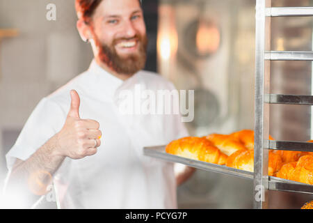 Un giovane bello baker mostra il suo pollice in alto con un foglio di cornetti freschi nelle sue mani contro lo sfondo di un forno. Foto Stock