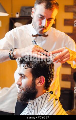 Barbiere cesoie capelli sulla sua testa con un giovane bel ragazzo con la barba e baffi. Barbiere. Foto Stock