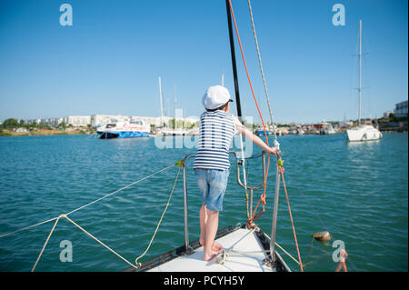 Un sano piccolo ragazzo in captain hat in piedi su yacht di lusso board nel porto di mare in estate Foto Stock