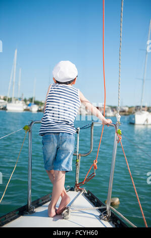 Little Boy caucasica nel capitano hat in piedi sul bianco scheda yacht nel porto di mare in estate Foto Stock