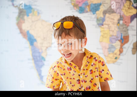 Emotional ridere caucasian capretto con occhiali da sole sulla testa sulla mappa del mondo sullo sfondo Foto Stock