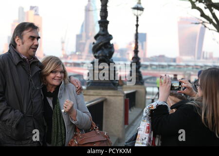 La mezza età matura in posa per una foto sulla riva del fiume Tamigi con la London City View in background, come il loro amico femmina prende foto sullo smartphone. Foto Stock