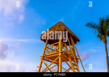 Giallo con tetto in paglia e legno torre bagnino o stazione torri oltre l'oceano dei Caraibi contro una vibrante tropical blu cielo con Palm tree Foto Stock