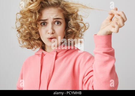 Studio shot di sconvolgere interrogato curly-dai capelli donna bionda tirando ciocca di capelli che sono rimasti subito dopo frizzle, essendo insoddisfatto del servizio con lei Foto Stock