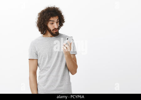 Dopo lungo tempo ex-girlfriend chiamando. Sorpreso storditi emotiva di ragazzo con i capelli ricci e la barba, fissando lo schermo dello smartphone con espressione stupita, l Foto Stock