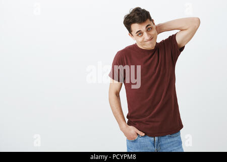 Piscina colpo di colpevole in imbarazzo europeo modello maschile in casuale t-shirt rossa, graffiatura retro del collo e guardando da sotto la fronte con le labbra aspirato, Foto Stock