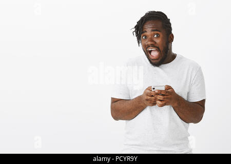 Ritratto di divertente Emotivo afro-americano modello maschio in abito casual, tenendo lo smartphone e la caduta della mascella, urlando da emozione e sorpresa wh Foto Stock