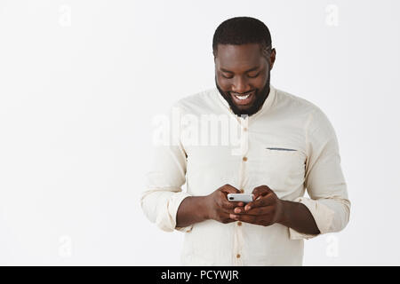 Guy godendo nuovo gioco, essendo intrattenuti e divertiti con le nuove funzioni dello smartphone. Ritratto di bella gioiosa americano africano in bianco alla moda di shi Foto Stock