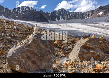 Un piccolo pupazzo di neve su una pietra in alta montagna contro lo sfondo di un ghiacciaio, picchi di montagna e un cielo blu in estate Foto Stock