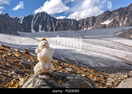 Un piccolo pupazzo di neve su una pietra in alta montagna contro lo sfondo di un ghiacciaio, picchi di montagna e un cielo blu in estate Foto Stock