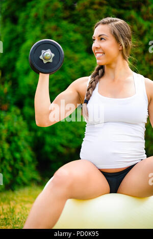 Donna incinta sorridente durante il sollevamento di pesi sulla sfera di Yoga Foto Stock