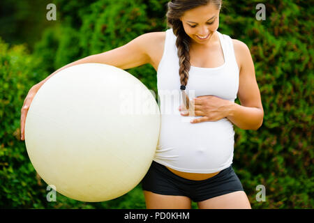 Donna incinta toccando ventre tenendo palla Fitness in posizione di parcheggio Foto Stock