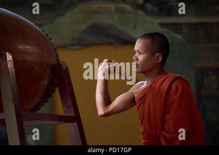 Un giovane monaco buddista giocando un tamburo durante una cerimonia in un monastero a Siem Reap, Cambogia, con facce di divinità dipinta in un murale in backgrou Foto Stock