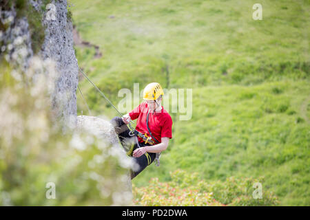 Foto di atleta ragazzo nel casco clambering over rock contro lo sfondo di verdi alberi Foto Stock