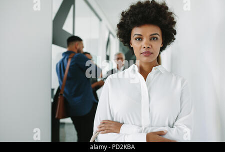 Ritratto di fiducioso imprenditrice africana in piedi in ufficio corridoio con le braccia incrociate e il suo team di incontro in background. Donna in office corri Foto Stock