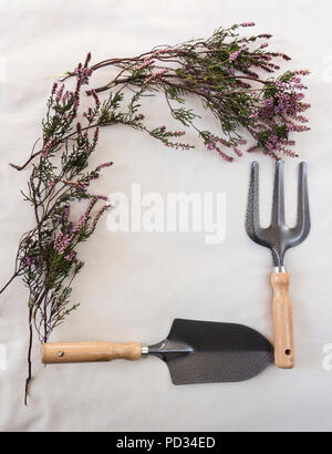 Viola autunno erica (Erica) telaio con attrezzi da giardino set - Forcella a mano a mano e frattazzo su ecru pezzo grezzo tessuto di cotone. Foto Stock