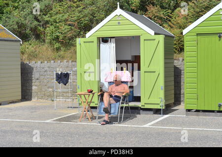 Uomo seduto su una sedia che legge fuori di una capanna verde spiaggia. Bournemouth, Dorset, Regno Unito, lunedì 6th agosto 2018. Il tempo caldo sulla spiaggia della costa meridionale mentre la gloriosa ondata di caldo estiva continua. Foto Stock