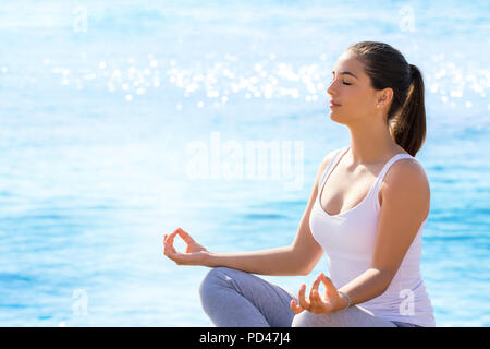 Close up ritratto di giovane donna meditando in posizione di yoga all'aperto. Ragazza in abbigliamento casual seduto contro il mare blu sfondo con sun riflettendo su Foto Stock