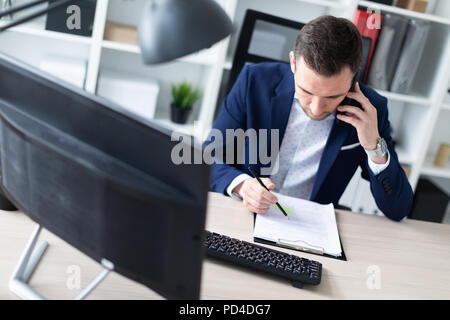 Un giovane uomo è seduto in ufficio al tavolo, parlando al telefono e tenere una matita in mano. Il giovane uomo ha documenti. Foto Stock