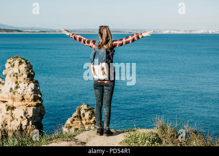 Una ragazza di turista o viaggiatore con lo zaino si ammira la splendida vista dell'Oceano Atlantico in Portogallo e solleva le mani in alto mostra come le piace e come essa è libera. Foto Stock