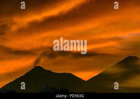 Fiery zampilli di lava dal vulcano Fuego nell'altipiano guatemalteco contro il fuoco del tramonto sfumature di colore come il fumo nuvole di cenere si mescolano con la sfocatura di nuvole. Foto Stock