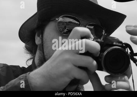 Ritratto in bianco e nero di un fotografo tenendo la sua fotocamera digitale, aviatore da indossare occhiali da sole e cappello del paese. Torbay, Airshow 2017. Devon, Regno Unito. Foto Stock