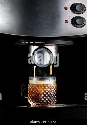 Drammatico colpo di una macchina per caffè espresso, closeup, infuso di caffè fresco in un bicchiere di vetro, con fumo di vapore in uscita Foto Stock