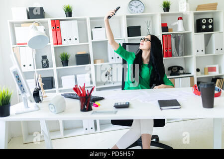 Una giovane ragazza con gli occhiali si siede in ufficio a tavola e scatta foto di se stessa sul telefono. Foto Stock