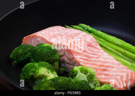 Filetto di salmone cotto a vapore con i broccoli e gli asparagi in una padella su uno sfondo nero, vista dall'alto Foto Stock