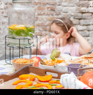 Una bambina mangia una torta e beve limonata. Cena in famiglia nel cortile all'aperto. Verdure e frutta autunnali sulla tavola. Tavolo da pranzo . Un luogo speciale per il testo Foto Stock