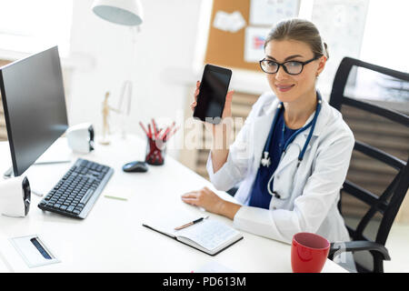 Una giovane ragazza in un abito bianco si siede a un tavolo in ufficio ed è in possesso di un telefono cellulare in mano. Uno stetoscopio appeso attorno al collo. Foto Stock