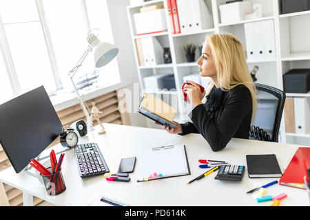 Una giovane ragazza è seduta al tavolo in ufficio, tenendo in mano un bicchiere e la lettura di un libro. Foto Stock