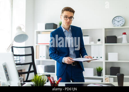 Un giovane uomo sorge nei pressi di un tavolo in ufficio e conserva documenti nelle sue mani. Foto Stock