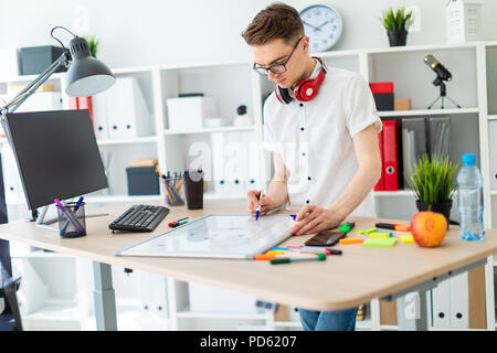 Un giovane uomo in bicchieri sorge vicino a un computer desk. Un giovane uomo estrae un indicatore su una scheda magnetica. Sul collo, il ragazzo di appendere le cuffie. Foto Stock