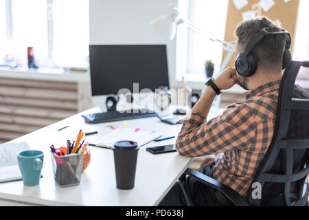 Un giovane uomo nelle cuffie si siede a un tavolo in ufficio e guarda il  monitor. La foto è stata scattata da dietro la spalla Foto stock - Alamy
