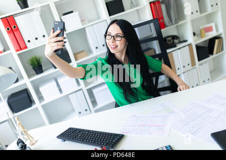 Una giovane ragazza con gli occhiali si siede in ufficio a tavola e scatta foto di se stessa sul telefono. Foto Stock