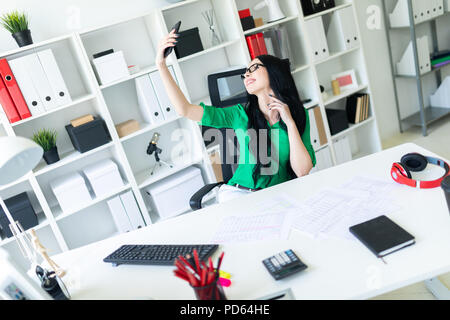 Una giovane ragazza con gli occhiali si siede in ufficio a tavola e scatta foto di se stessa sul telefono. Foto Stock