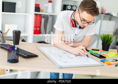 Un giovane uomo in bicchieri sorge vicino a un computer desk. Un giovane uomo estrae un indicatore su una scheda magnetica. Sul collo, il ragazzo di appendere le cuffie. Foto Stock