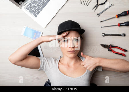 In grado giovane donna salutando la telecamera e dando un pollice in alto come lei giace sul pavimento circondato dai suoi dispositivi digitali e gli strumenti per la riparazione e mai Foto Stock